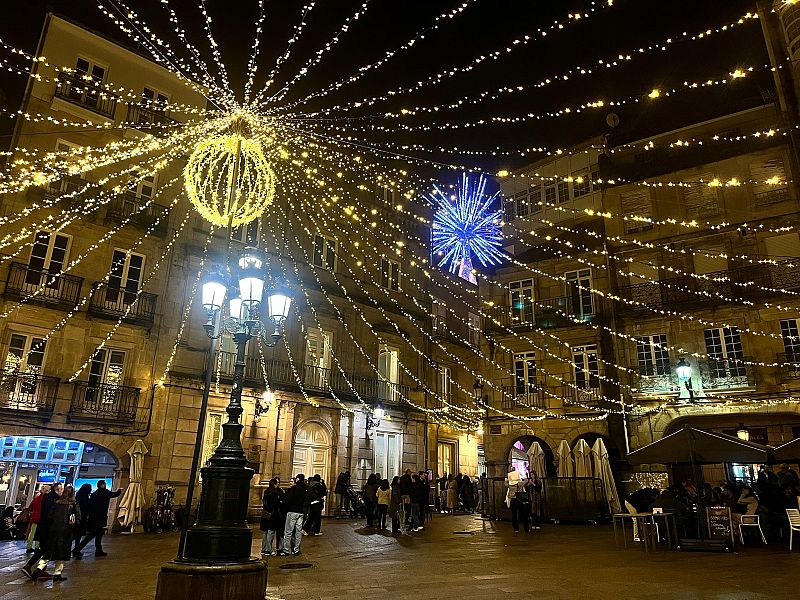 Una plaza española se ilumina con motivos navideños, incluyendo una esfera amarilla y una estrella azul. La escena nocturna muestra edificios clásicos y personas disfrutando del ambiente festivo.