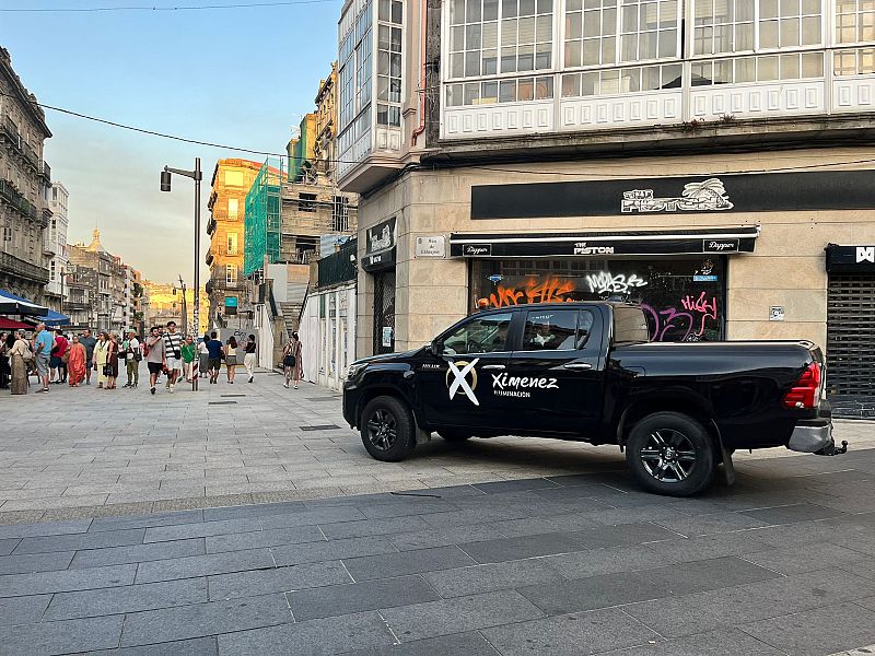 Una calle española adoquinada es el escenario donde se observa una camioneta negra Toyota estacionada, con el logo de Ximenez Iluminación. Al fondo, se distinguen locales comerciales y edificios antiguos.