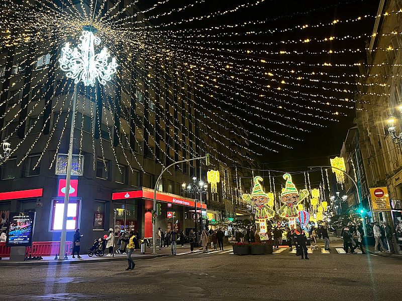 Una calle iluminada por luces navideñas en España, con adornos colgantes y figuras festivas. Se distingue la palabra "VIGO" en uno de los adornos y un edificio bancario en la esquina.