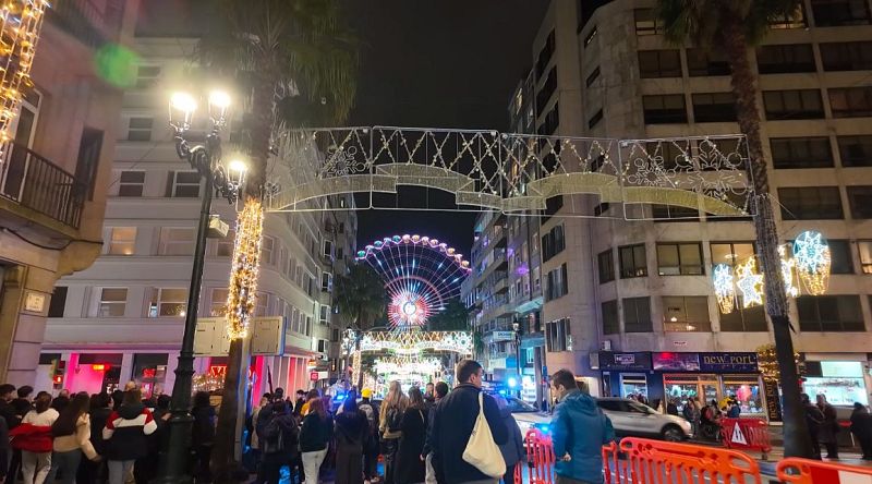 Una vista nocturna de una calle decorada con luces navideñas muestra una noria iluminada al fondo. La calle, llena de gente vestida con ropa de abrigo, transmite un ambiente festivo y alegre.