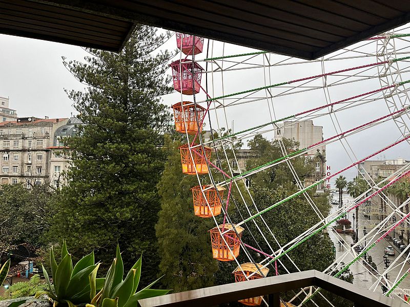 Desde el interior de una vivienda, se observa una gran noria con cabinas de colores, ubicada frente a edificios clásicos y vegetación. El cielo gris y la presencia de una barandilla indican que la fotografía fue tomada desde un balcón.