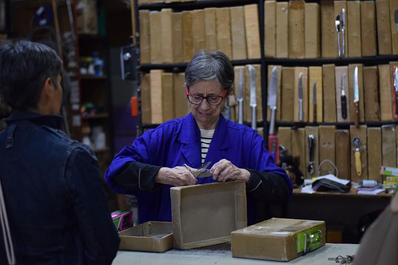 En una tienda de Madrid, Pepa Molina, con gafas rojas y bata azul, corta una caja de cartón. Un cliente con chaqueta vaquera está de espaldas, y sobre la mesa hay más cajas. Detrás, una estantería con utensilios.