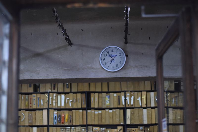 El interior de una ferretería antigua se revela con estanterías repletas de herramientas y cajas de madera, un reloj de pared y cadenas metálicas, creando una atmósfera nostálgica.