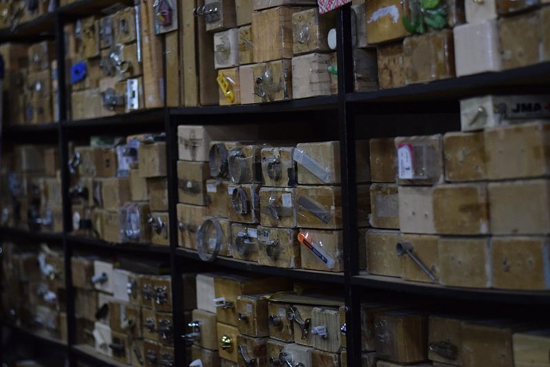 En la imagen se observan estanterías repletas de cajas de madera con objetos metálicos visibles, como cerraduras y llaves, sugiriendo un entorno de almacenamiento o taller.