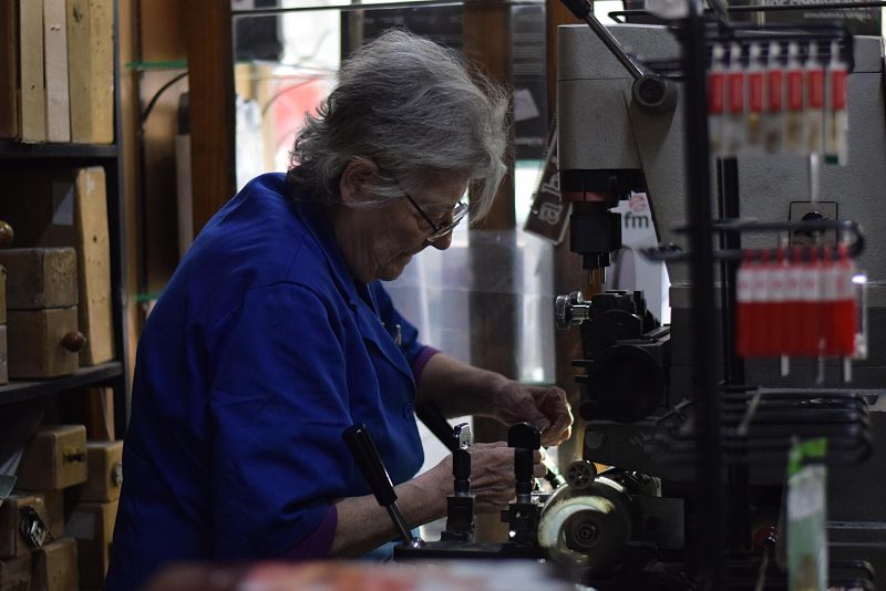 Alicia Molina, con gafas y cabello gris, trabaja en su taller madrileño. Se enfoca en copiar llaves con una máquina antigua, vestida con una camisa azul.