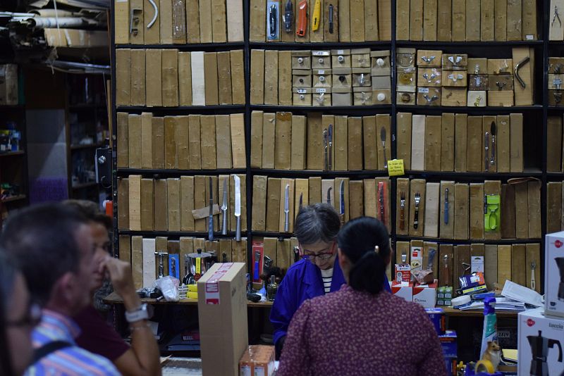 En una tienda, Pepa Molina, con gafas y bata azul, atiende a clientes. Se observan cajas de madera con utensilios y clientes, incluyendo una mujer con blusa floral y un hombre con camisa a cuadros.