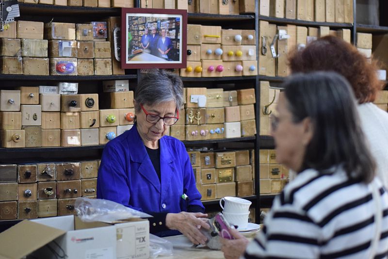 En una ferretería, una mujer mayor con gafas y chaqueta azul atiende a dos clientas. Detrás del mostrador, manipula un objeto mientras una clienta habla por teléfono y otra observa. Se aprecian estanterías con cajones de madera y una foto en la parte superior.