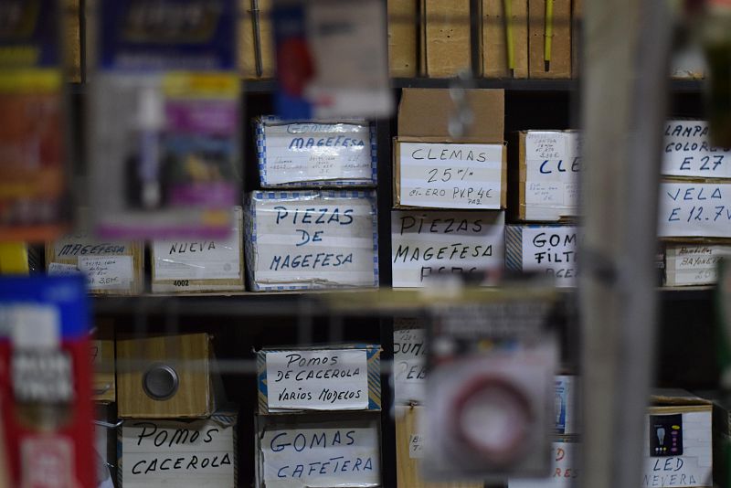 La imagen muestra una estantería llena de cajas de cartón con etiquetas escritas a mano, identificando productos como gomas, piezas, pomos y repuestos de iluminación.