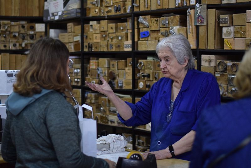 María Molina, con mono azul y gafas, atiende a un cliente en una ferretería. El cliente, de espaldas, lleva una chaqueta gris y una bolsa blanca. El fondo muestra estanterías llenas de cajas de madera con objetos.