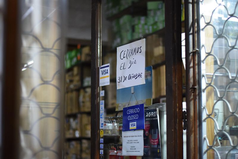 En la entrada de una ferretería, se exhiben carteles que informan sobre el cierre del establecimiento el 31 de octubre y su estado como "Cerrado". Se aprecia un logo de Visa y una puerta con reja metálica.