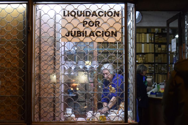 En el interior de una ferretería, un hombre mayor se concentra en organizar objetos, mientras una persiana metálica a medio bajar anuncia una "Liquidación por Jubilación".