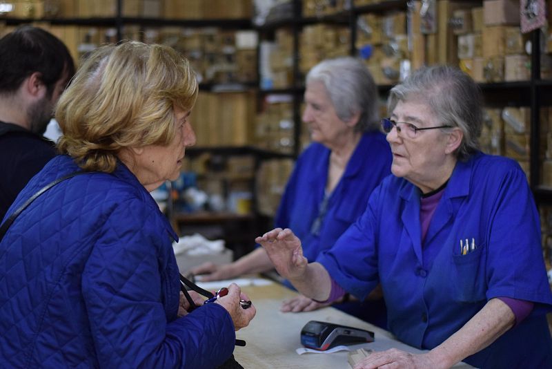 Dos mujeres, identificadas como María y Alicia, atienden en una ferretería. Alicia, con chaqueta azul y gafas, habla con María, que lleva uniforme de trabajo y atiende a un cliente. En el mostrador hay un terminal de pago.