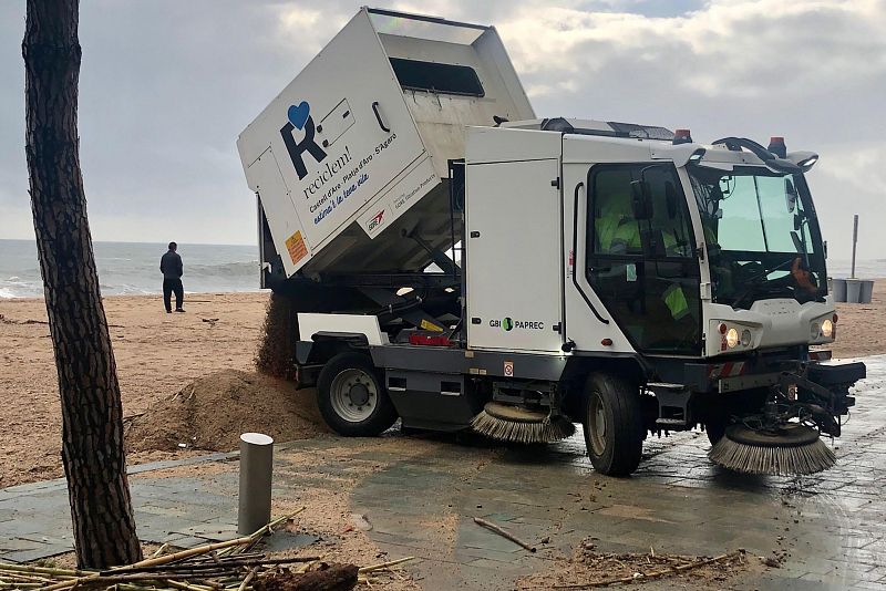 Una máquina blanca limpia arena de un paseo marítimo, con un contenedor que muestra texto. Se observa una playa con arena y el mar al fondo, y una persona de pie.