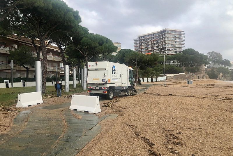 Una playa con poca arena y restos de vegetación tras una tormenta. Un camino bloqueado por barreras de plástico conduce a la playa, donde un vehículo de limpieza está estacionado. Un hombre observa la escena con el cielo nublado.