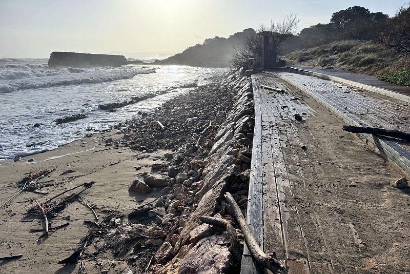 En una playa soleada, se aprecia arena y rocas en primer plano, con olas rompiendo en la orilla. Un paseo marítimo de madera, cubierto de arena, se extiende a la derecha, indicando posiblemente trabajos de limpieza.