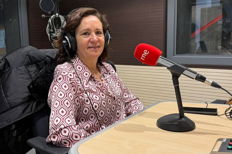 Isabel Bartolomé, con auriculares y camisa estampada, está frente a un micrófono rojo en un estudio de radio. Se encuentra sentada frente a una mesa de madera clara.
