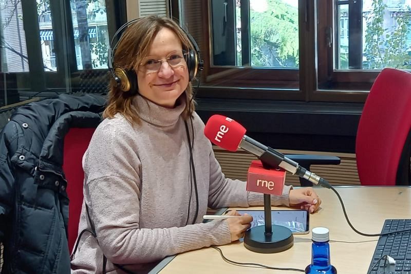 Mar Rubio-Varas, con gafas y auriculares dorados, está en un estudio de radio frente a un micrófono rojo con el logo de RNE. Viste un jersey de cuello alto claro y hay una tableta y una botella de agua azul sobre la mesa.