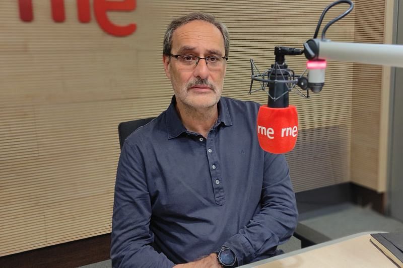 Francisco Cayón, profesor, aparece en un estudio de radio, con un micrófono de RNE detrás. Viste una camisa oscura y lleva gafas, bigote y pelo canoso.
