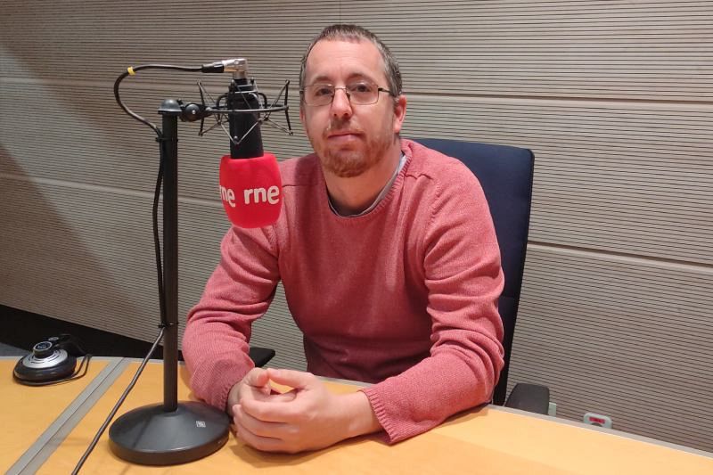 José Luis Velasco, con gafas y barba, se encuentra en un estudio de radio frente a un micrófono con el logo de la emisora. Viste un jersey rosa y está sentado delante de una pared con paneles.