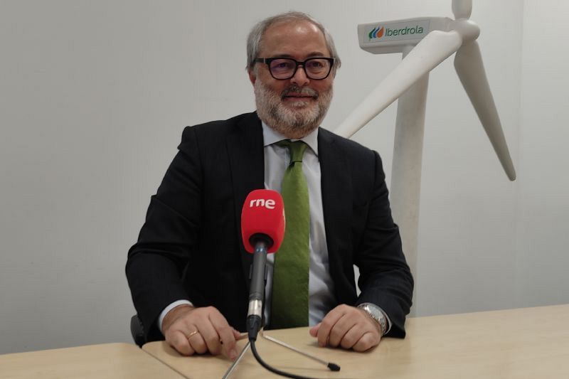 Julio Castro, directivo de Iberdrola, aparece sentado frente a un micrófono con el logo de RNE, vestido con traje y corbata. Al fondo, se distingue una maqueta de una turbina eólica.