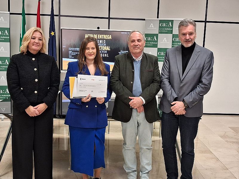 María Teresa Donstrup, con un traje azul, recibe un premio en un evento formal. La acompañan otras tres personas, dos hombres y una mujer, en un escenario con banderas y una pantalla con texto relacionado con un premio audiovisual.