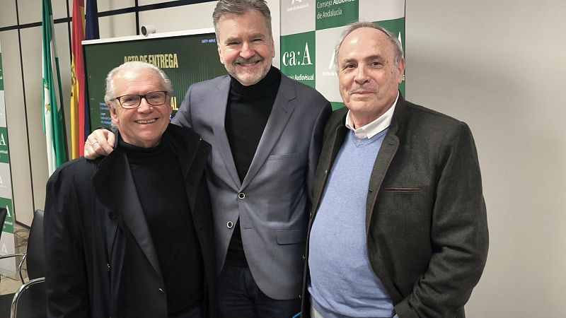 Tres hombres posan para una foto en un evento. Uno lleva un jersey negro de cuello alto y abrigo, otro un jersey negro y americana gris, y el tercero una camisa blanca, jersey azul y chaqueta marrón. Detrás, un panel con un logotipo y banderas.