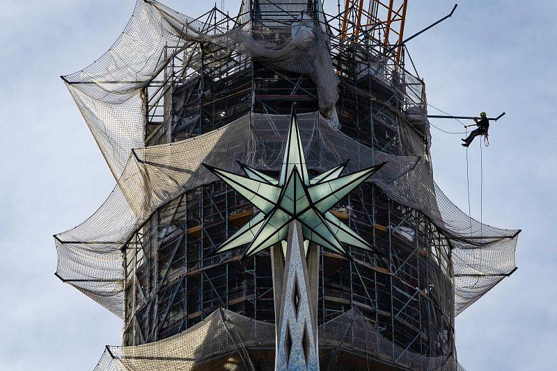 En la imagen se aprecia una torre en construcción, cubierta por andamios y redes de seguridad. En la parte superior, se distingue una cruz con forma de estrella. Una persona se encuentra trabajando en altura, utilizando cuerdas.
