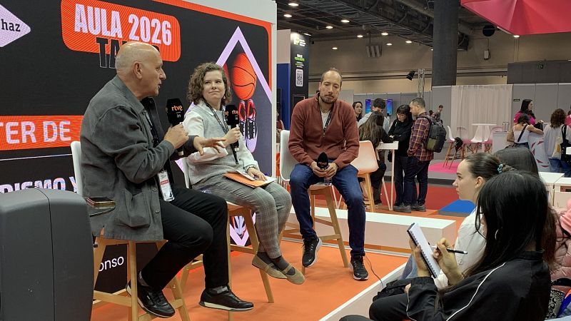 Tres ponentes, con micrófonos, participan en un taller. Un hombre calvo con chaqueta gris, una mujer con camisa blanca y chaleco gris, y otro hombre con camisa marrón rojiza y vaqueros, se encuentran frente a un panel con texto y elementos gráficos.