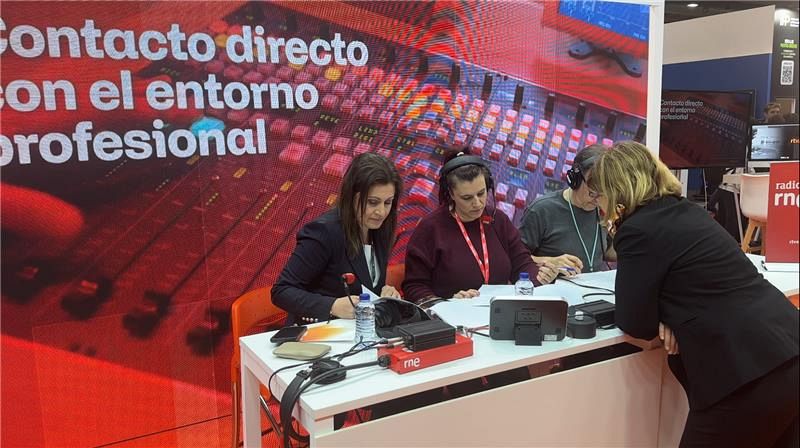 Tres mujeres en un estudio de radio, posiblemente de RNE, interactúan frente a micrófonos y una pantalla con texto. Dos están sentadas y una de pie, todas vestidas con ropa casual.