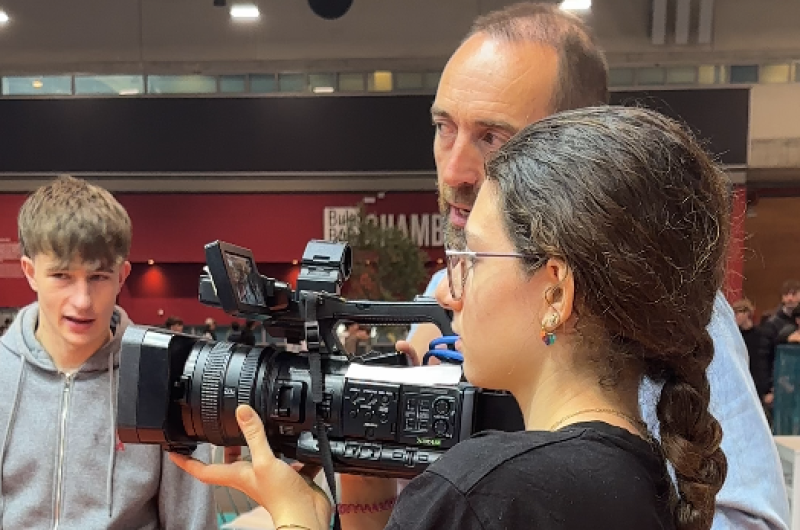 Una mujer con gafas y trenza maneja una cámara de televisión negra con un gran objetivo, posiblemente en un taller. Un hombre y un joven la acompañan.