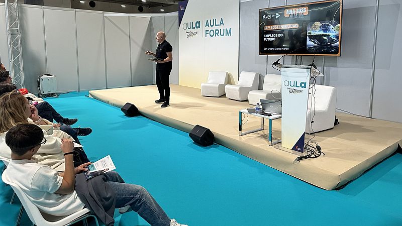 Un hombre calvo, vestido de negro, imparte una charla sobre IA y sostenibilidad en un evento. En el fondo, se aprecia el logo de RTVE y el título de la ponencia, mientras el orador gesticula con una tablet.