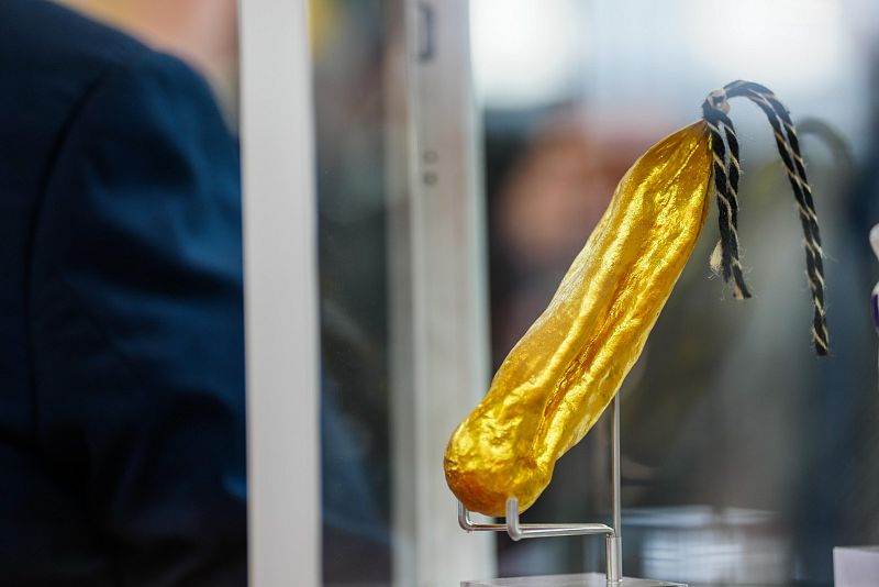 Un embutido de forma cilíndrica y recubrimiento dorado, posiblemente un fuet o salchichón, se exhibe en una vitrina de cristal. Se aprecia una cuerda negra y dorada en uno de sus extremos, y está colocado sobre un soporte metálico.
