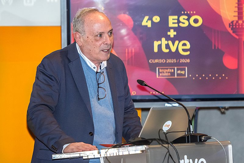 Esteban Mayoral, con gafas y vestimenta azul, se encuentra frente a un atril con un ordenador portátil Panasonic y un micrófono. Al fondo, una pantalla con el logo de RTVE y texto sobre un curso.