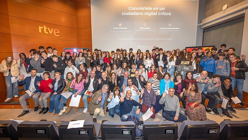 En un auditorio, un grupo de estudiantes de secundaria posa para una foto junto a adultos, todos mostrando cuatro dedos. Una pantalla al fondo proyecta un mensaje sobre la importancia de la ciudadanía digital.