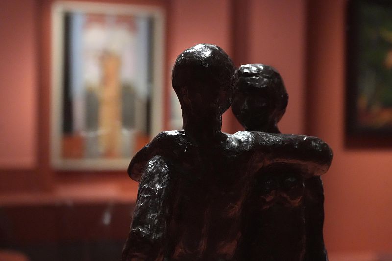 Una escultura oscura de dos figuras abrazadas, con textura rugosa, se destaca en primer plano. Al fondo, se vislumbran dos cuadros borrosos sobre una pared de color rojizo.