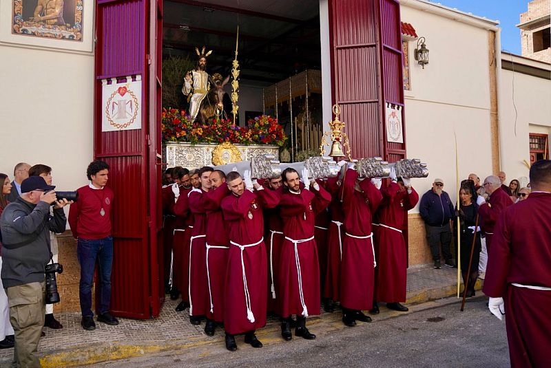 En una calle de Melilla, una procesión religiosa avanza con un paso adornado y una imagen de Jesús montado en un burro, transportado por hombres con túnicas burdeos.