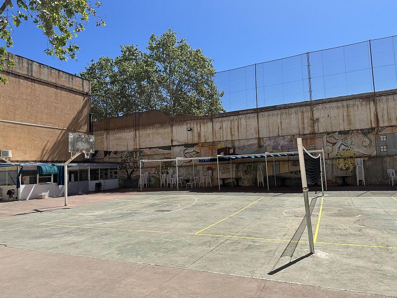 Una pista deportiva exterior con una red de voleibol en el centro, una canasta de baloncesto en un lateral y un edificio al fondo. El cielo es azul y hay árboles que sobresalen por encima de una pared.
