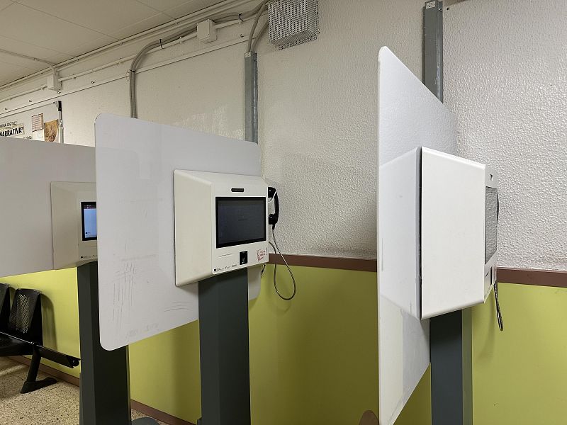 Se observan varias cabinas individuales para videollamadas, equipadas con pantallas, teléfonos y paneles protectores blancos. El entorno presenta paredes claras con una sección inferior amarilla y sillas negras en el fondo, sugiriendo un área de espera.