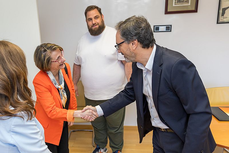 José Luis Pérez Pastor (RTVE) y Eva Alcón Soler (UJI) estrechan manos en un encuentro profesional. Un hombre con barba y camiseta blanca observa la escena.