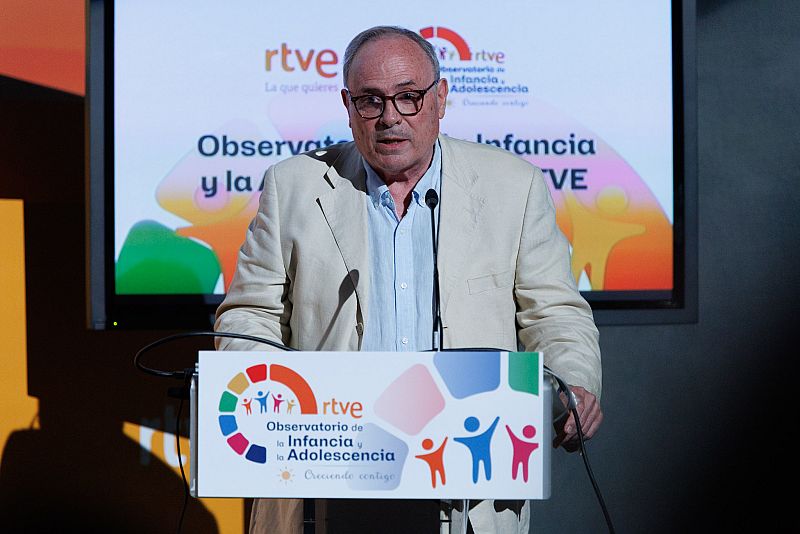 Esteban Mayoral habla detrás de un atril con logos de RTVE y "Observatorio de la Infancia y la Adolescencia", lema "Creciendo contigo", y siluetas de niños y personas en pantalla.