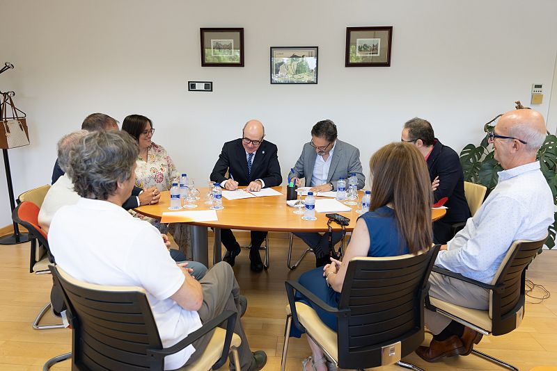 José Manuel Pérez Tornero y un representante de la Universidad de Granada firman documentos en una reunión formal. Ocho personas observan el acto en una sala de juntas con decoración y elementos de oficina.