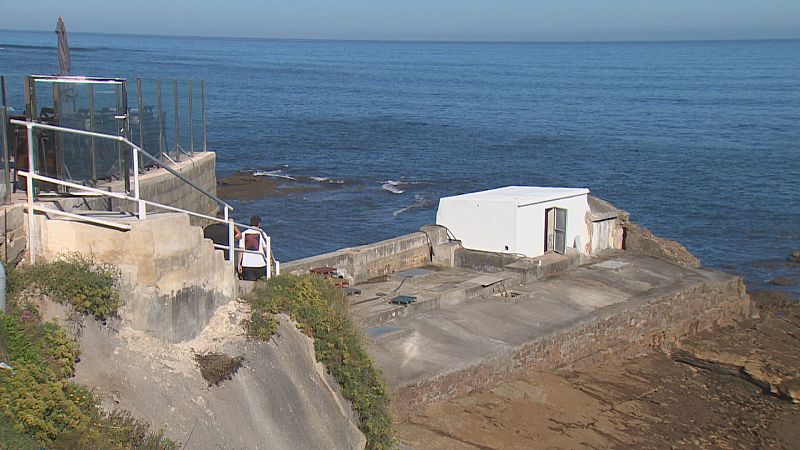 Paisaje costero con edificaciones de hormigón en una ladera rocosa, mar azul y una persona descendiendo escaleras. Se observa un edificio blanco sobre una plataforma.