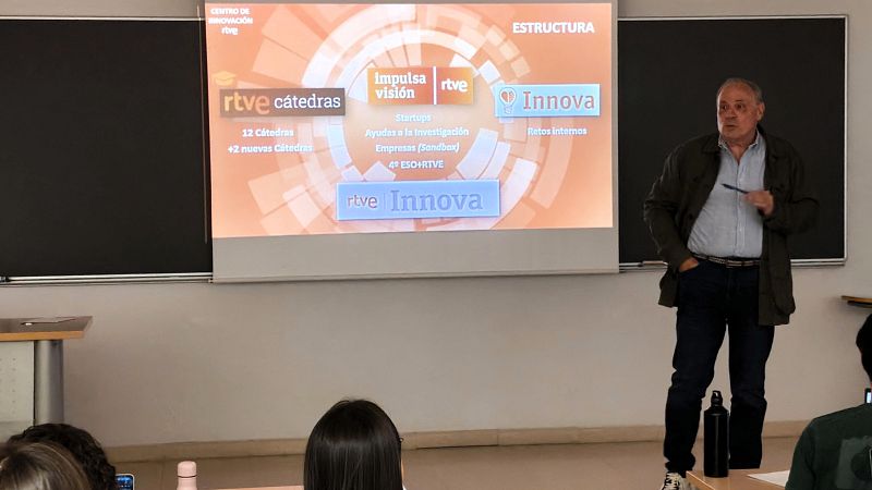 Esteban Mayoral presenta la estructura del Centro de Innovación de RTVE ante un público, apoyado por una diapositiva proyectada en la pared de un aula.