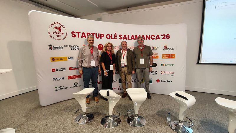 Cuatro personas con acreditaciones posan ante un panel corporativo blanco con letras rojas y logotipos de patrocinadores.