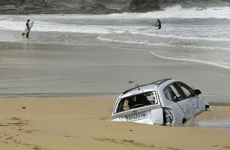 Dos surfistas se prepraran para entrar en el agua frente a un coche hundido en la arena de la playa de Verdicio, Asturias.