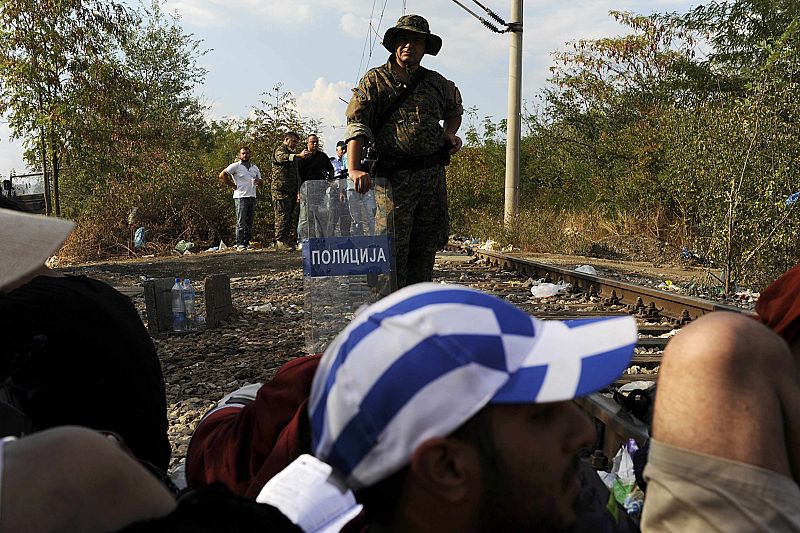 Macedonian police officer stands guard on the borderline with Greece as migrants rest on the Greek side of the borders