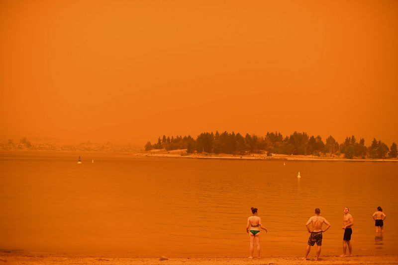Un grupo de personas se refresca en el lago Jindabyne, bajo un cielo anaranjado debido al humo procedente de los incendios