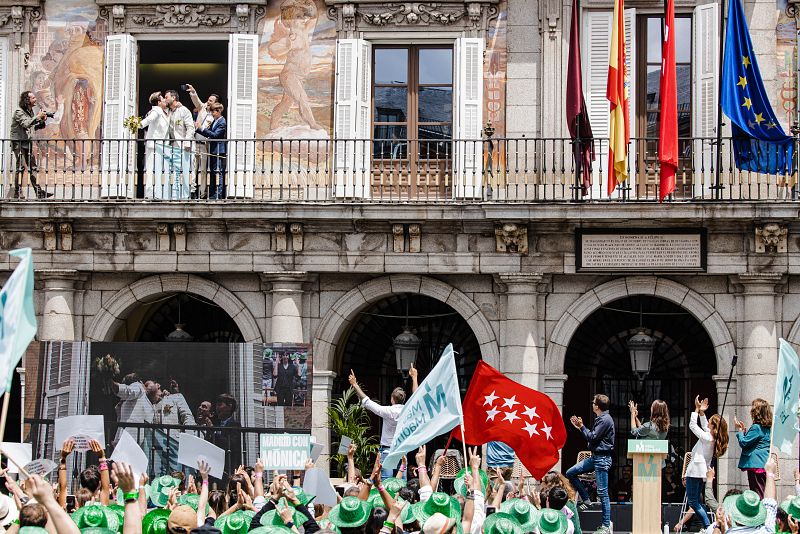 Unos recién casados sacan una bandera de Más Madrid en el balcón durante un mitin del partido