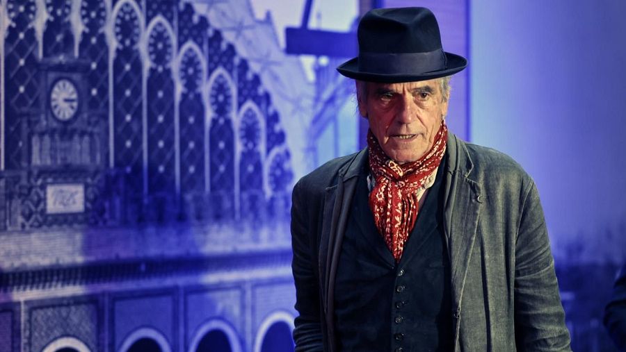 El actor británico Jeremy Irons, con sombrero y pañuelo rojo, en una presentación o entrevista para "De película" de RNE.  Se ve una imagen borrosa de un edificio al fondo.
