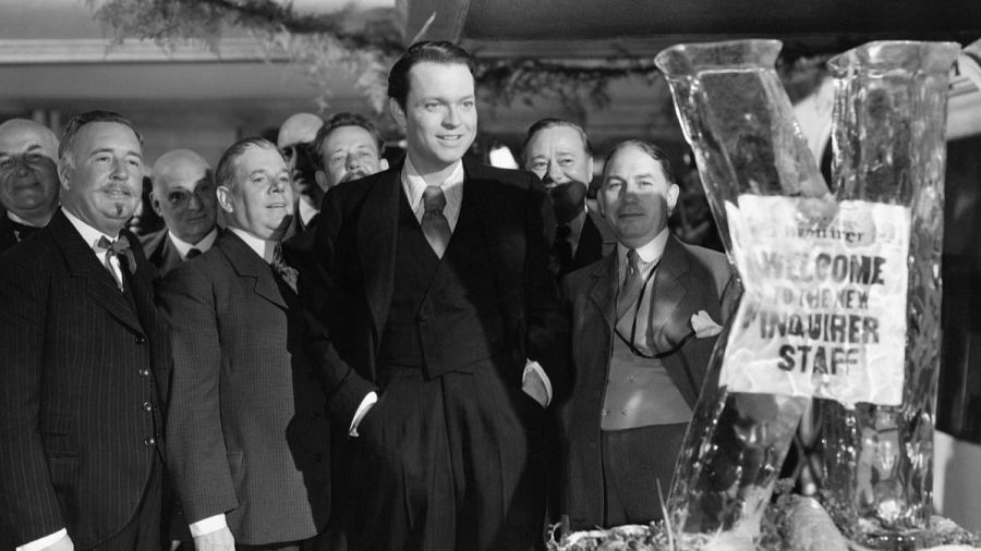 Orson Welles, en el centro, rodeado del equipo del periódico Inquirer en una escena de "Ciudadano Kane". Una escultura de hielo da la bienvenida al nuevo personal. No se identifican personajes públicos españoles.
